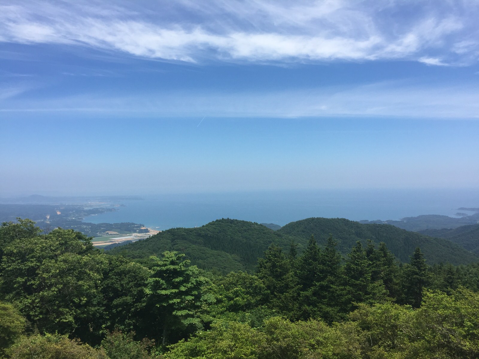 緑豊かな山々の向こうに青い海が広がり、晴れた空に薄い雲が浮かんでいる景色の写真