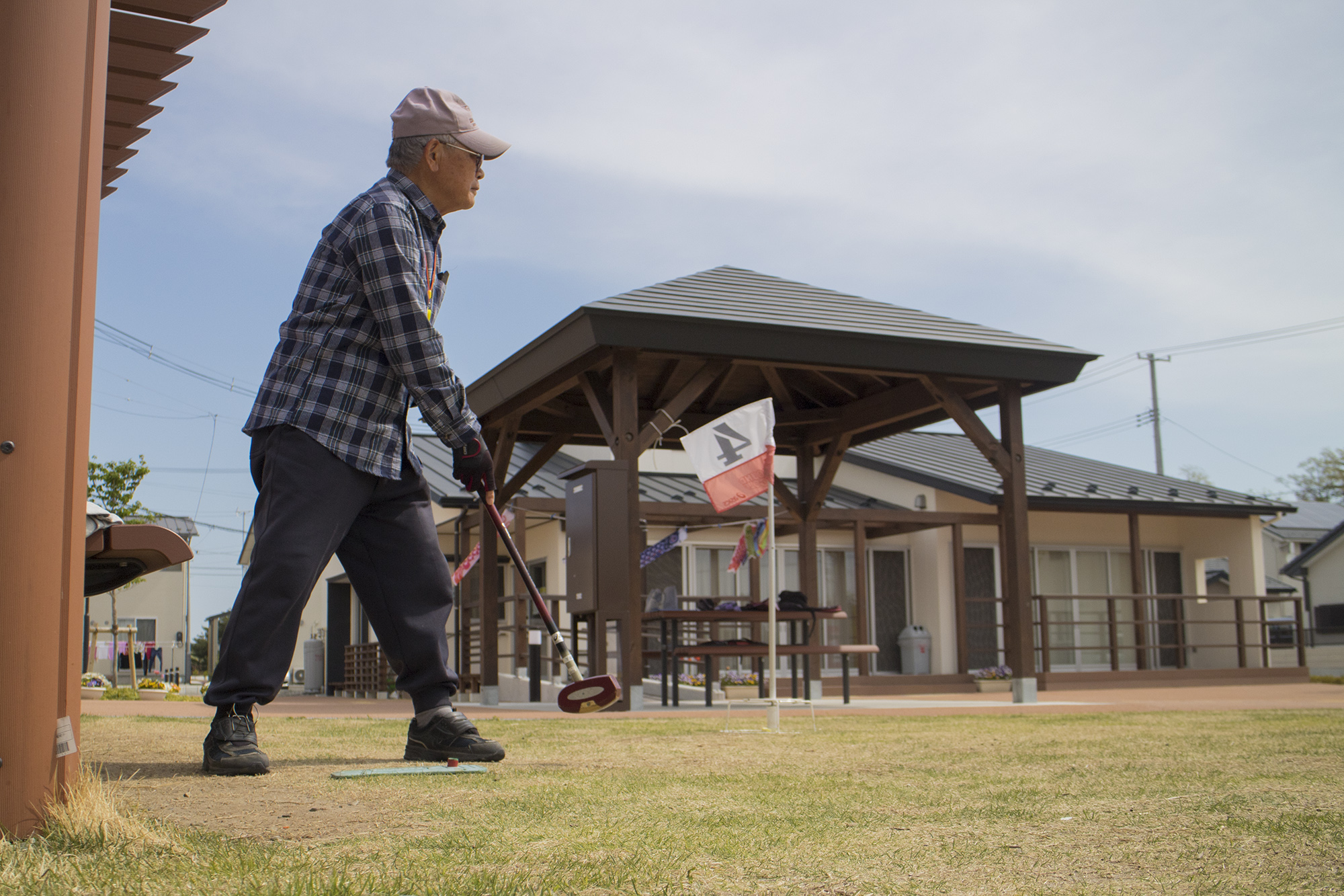 屋外の芝生の上で、キャップとチェック柄のシャツを着た高齢の男性が、グラウンドゴルフ用のスティックを構え、打ったボールの方を見つめている様子の写真