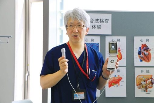 青い医療用スクラブを着た白髪の住友和弘医師が、手にポータブル型の超音波検査機器を持ちながら説明している様子の写真