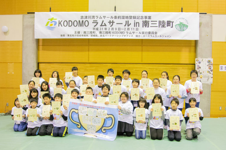 「KODOMOラムサールin南三陸町」と書かれた横断幕が掲げられた室内で、優勝カップの様なイラストが描かれた大きな紙を前に置き、一人ずつ黄色の用紙を持った子どもたちが並んでいる写真