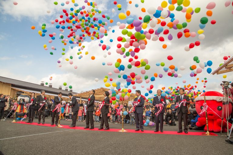 沢山の風船が上空に舞い上がり、ねじり鉢巻きをしたオクトパス君や園児たちが見守るなか、レッドカーペットの上に胸章リボンをつけた関係者が横一列に並び、テープカットを行っている様子の写真