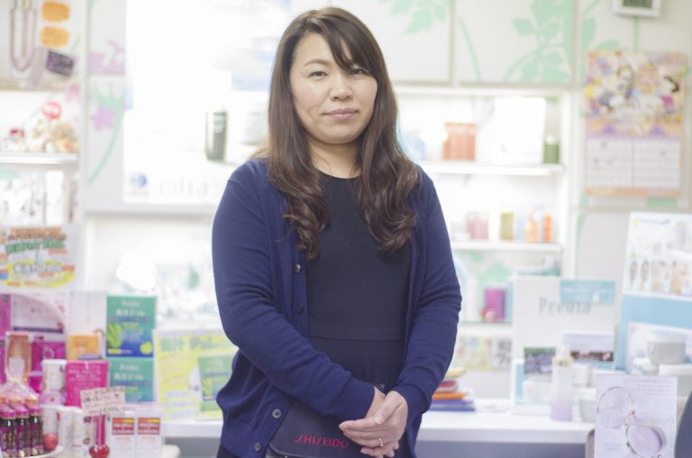 棚や机の上に沢山の化粧品が並んでいる前に立つ、紺色のカーディガンを着た佐藤さんの写真
