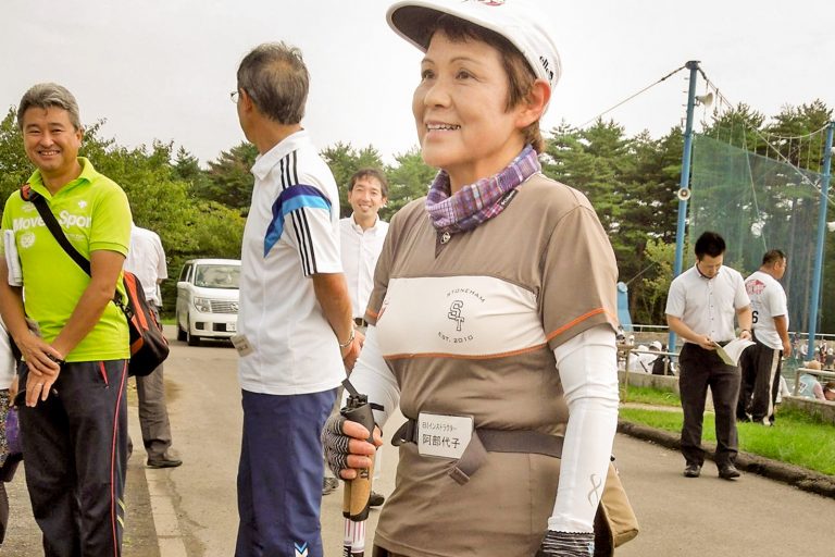 背後に複数の人々が立っている中、駐車場で白いキャップを被り、腕カバーをつけ、ベージュ色のシャツを着た阿部さんが立っている写真