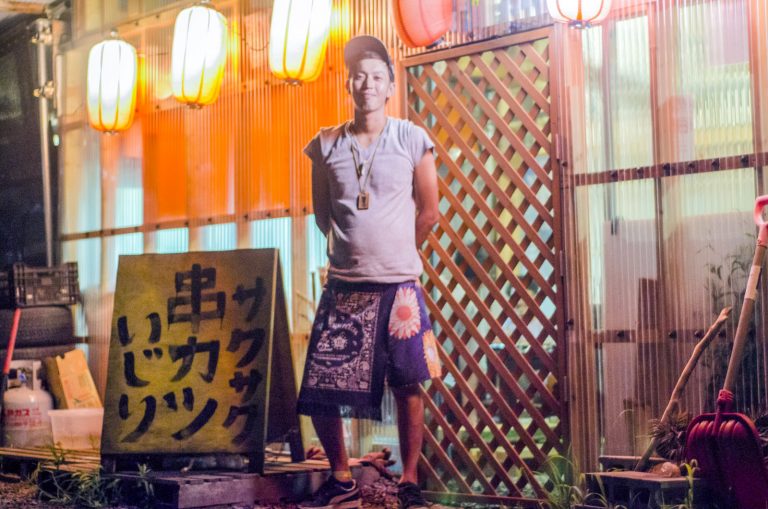 照明が灯された提灯が飾られたお店の前の「サクサク串カツいじり」と書かれた黄色の看板の傍に立つ、キャップを被りグレー色のシャツを着た井尻さんの写真