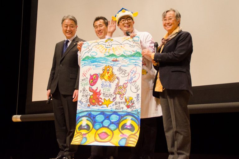 フグのアップのイラストや、南三陸のきれいな自然や魚介類のイラストが描かれた大きな模造紙を、3人の男性とフグの被り物を被ったさかなクンが横一列に並んで持ってステージに立ち、さかなクンが右手を上げているラムサール条約シンポジウムの様子の写真