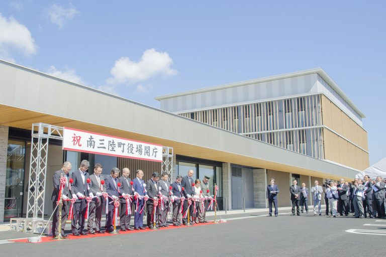 新しい建物の前に設置された「祝 南三陸町役場開庁」と書かれた横断幕の前で、スーツ姿の関係者の方々が、赤いリボンのついたテープカット用のテープをハサミで切っている新庁舎開庁の式典の様子