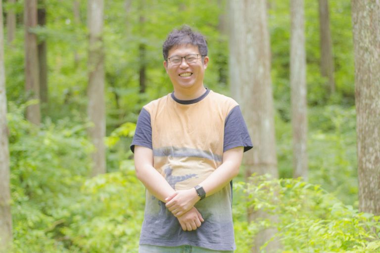 草が生い茂り、木々が密集した林の中に立つ、笑顔の佐藤さんの写真