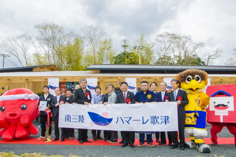 レッドカーペットの上にオクトパス君や他2体のマスコットキャラクターが両端に立ち、「南三陸ハマーレ歌津」と書かれた横断幕を持った、左胸に胸章リボンをつけた町長や関係者の方々が横一列に並んでいる写真