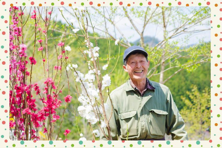 緑の木々に囲まれた山の上で、ピンクや白色の花が咲く木の傍に立つ、笑顔の緑の作業服と紺のキャップを被った男性をアップで撮影した写真