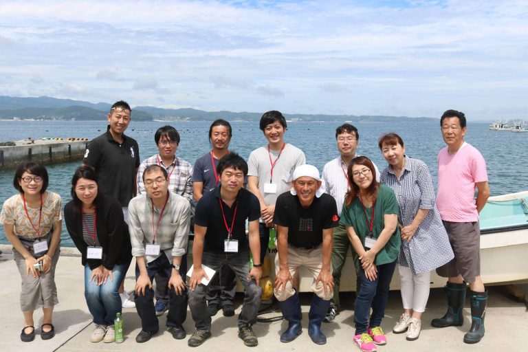 小型船が置かれた港の岸壁で、海を背景に名札を付けたツアー参加者が立ち並んでいる記念写真