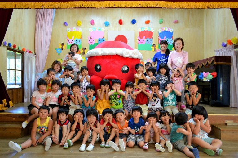 ピアノが設置された幼稚園の舞台で、大きな赤いねじり鉢巻きをしたオクトパス君を囲み、沢山の子どもたちと先生たちが笑顔でポーズを取っている記念写真