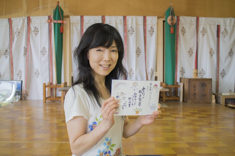 フローリング床の屋内で、黒髪の女性が笑顔でカメラを見つめながら、かわいらしいイラストが描かれた手書きの虹色アートの作品を両手で持っている様子をアップで撮影した写真