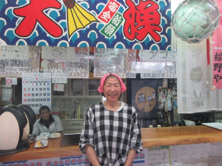 背景には色鮮やかな大漁旗やカレンダーが壁に飾られ、店内のカウンター越しにチェック柄の服を着た松野三枝子さんが笑顔で立っている写真