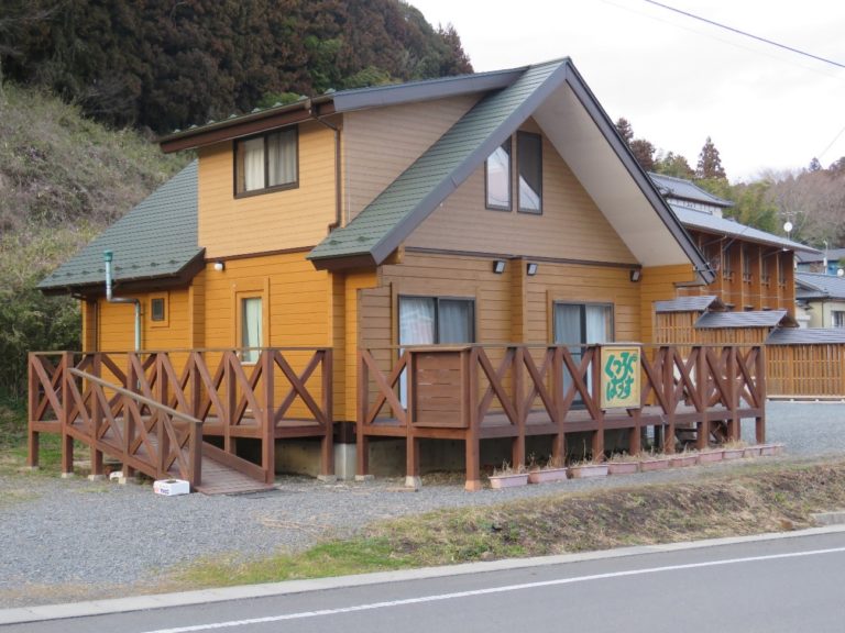 スロープや「くつろぐはうす」と書かれた看板が設置されたウッドデッキがある、道路沿いに建っている2階建ての木造のログハウスの建物外観の写真