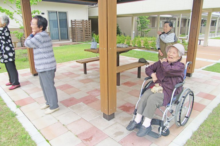 車椅子の女性を含む4人の女性が東屋の下で、左手を右肩に置いて体操をして楽しんでいる様子の写真