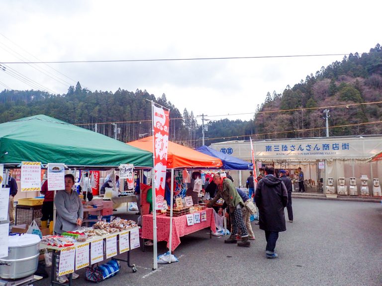山を背景に立つ南三陸さんさん商店街の前に、商品がたくさん並んだ緑、オレンジ、青の屋根のテントが立ち並び、「朝市」と書かれた赤いのぼりが立っているさんさん朝市が来場客でにぎわっている様子の写真