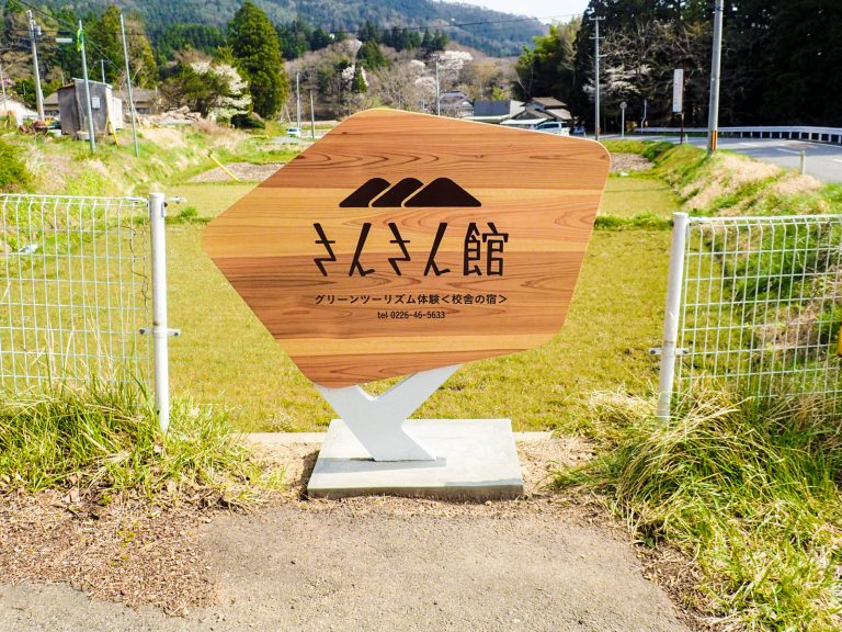 緑に囲まれた田舎の風景の中に「さんさん館」と書かれた木製看板が立てられている写真