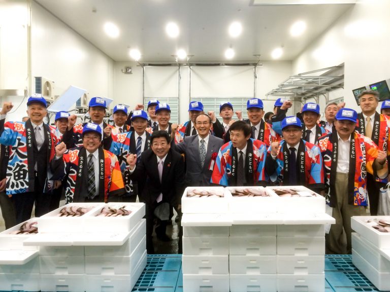 青い帽子と「大漁」と書かれたはっぴを着た関係者たちが、スーツ姿の町長と関係者の男性を中央に、積み重ねられた魚の入った白い発泡スチロール箱の前で笑顔で並んでいる落成記念の集合写真