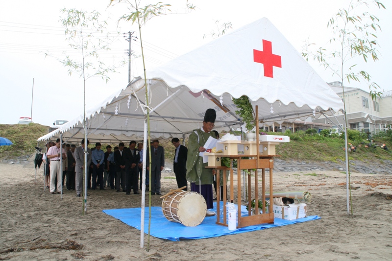 砂浜に設置されたテントの下に祭壇が設けられ、神職の男性が神事をおこなっており、多くの関係者の方々が参列し、安全祈願を行っている海開きの様子の写真