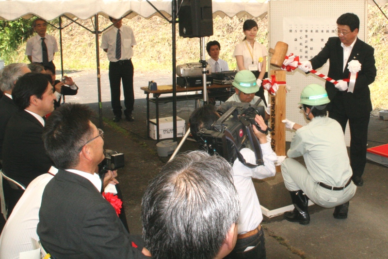 屋外のテントの下で、多くの関係者、報道陣が見守る中、紅白のリボンが巻かれた木槌を持った町長が、ヘルメットをかぶった2人の作業員の男性が支えている杭を打ち込もうとしている、三陸縦貫自動車道南三陸道路測量中心杭設置式の様子の写真