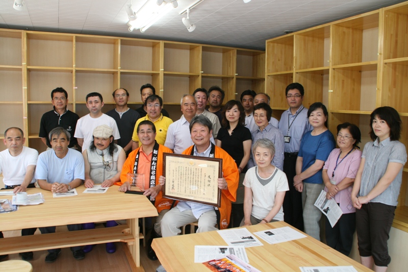 木製の棚が並ぶ室内で、多くの関係者の方々が集まり、オレンジ色の法被を着た前方中央に座る男性が賞状を掲げている受賞報告会の記念写真