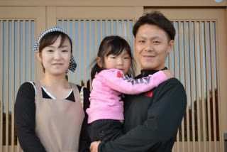 玄関の前で、笑顔の菅原さん夫婦とその子どもの娘さんご主人に抱っこされて並んで写っている写真