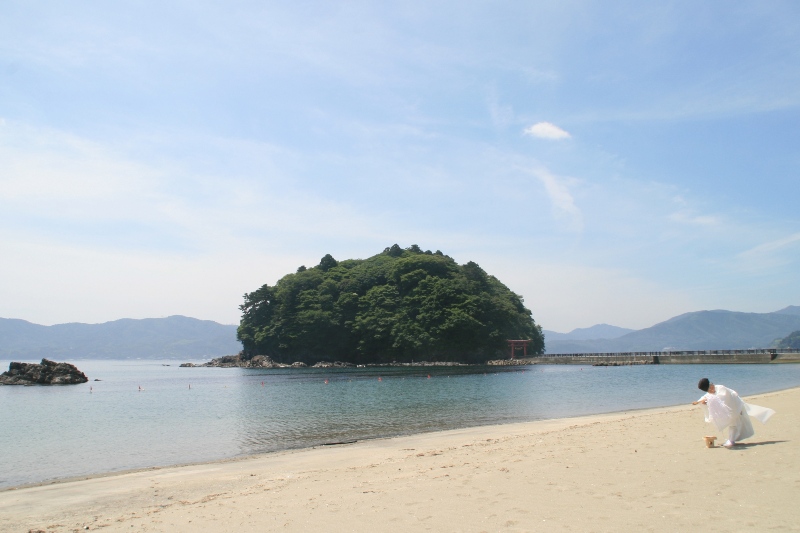 陸から伸びた道の先に赤い鳥居がある、緑豊かな小島が見える海辺の砂浜で、白い装束を着た神主が安全祈願を行っている海開きの様子の写真