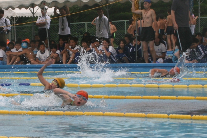 大勢の観客や応援の児童が見守る屋外プールで、ロープの張られたレーンを、ゴーグルと水泳帽を着用した選手たちが、力強いクロールで水を大きくかき分けて競い合っている水泳大会の臨場感あふれる写真