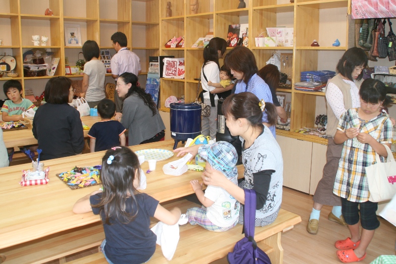 木製の棚に商品が並ぶ明るい店内に親子連れなど多数の客が集まっており、手前のテーブルでは親子連れが座って軽食をとったりして休憩している様子が写っているBOXショップ店内の様子を写した写真