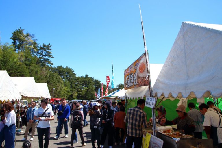 青空の下、白いテントが並ぶ屋外の祭り会場で、多くの人々が屋台の食べ物を買ったり見たりしているにぎやかなイベントの写真