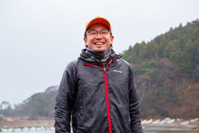 木々と岸辺に船や建物が遠目に見える水辺に立ち、オレンジ色のキャップをかぶり、黒いジャケットを着て笑顔の後藤伸弥さんの写真