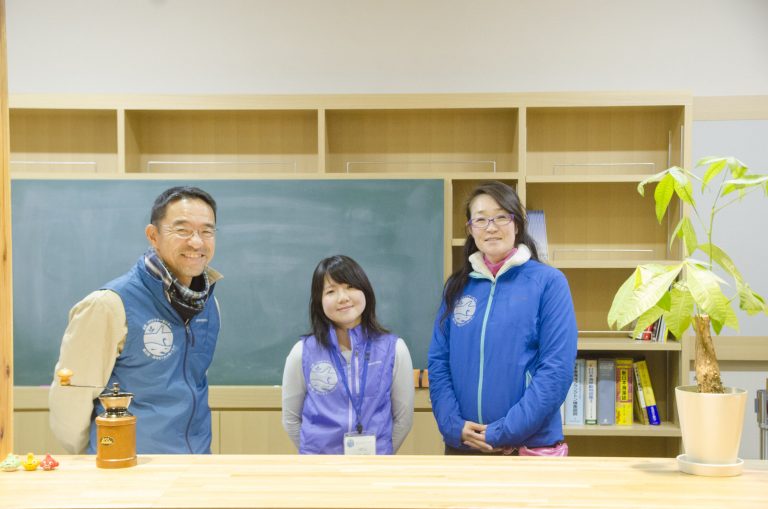 黒板が設置された棚の一部に書籍が並び、その前に右胸にお揃いのマークがついた色違いのジャケットを着た男性と2人の女性が並んで立っている写真