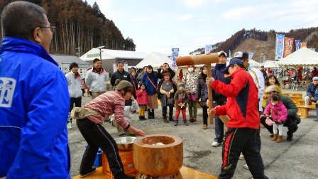 人々が見守る中、杵と臼を使ってもちつきをしている様子で、赤色の上着を着た男性がつき、ニット帽を被った女性が手を添えている写真