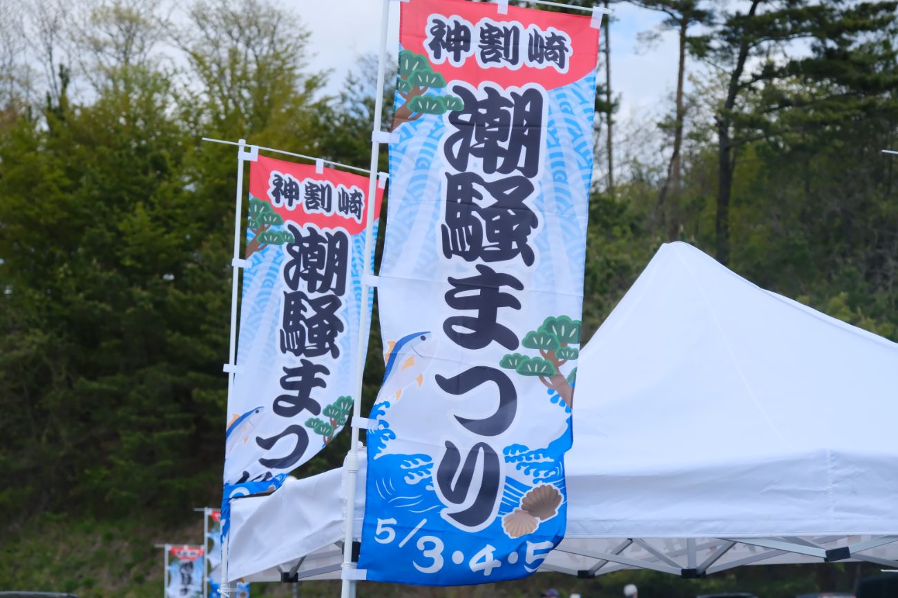 木々に囲まれたイベント会場で、白いテントが並ぶ販売ブースごとに設置された「神割崎潮騒まつり」と書かれたカラフルなのぼり旗が風になびいている様子の写真
