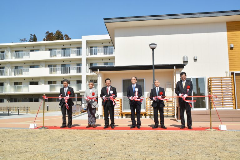 白壁の4階建ての団地の前にレッドカーペットが敷かれ、胸章リボンをつけた町長や5人の関係者が横一列に並びテープカットを行っている様子の写真