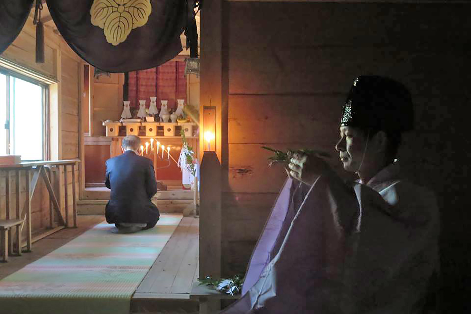神職の庄悦さんが暗がりの社殿の中で神前に向かって榊(さかき)を手に持ち祈りを捧げる中、スーツ姿の男性が正面の神棚に向かって正座し深く礼拝している様子が静かに灯されたロウソクの明かりに照らされている、神道の厳かな儀式の写真