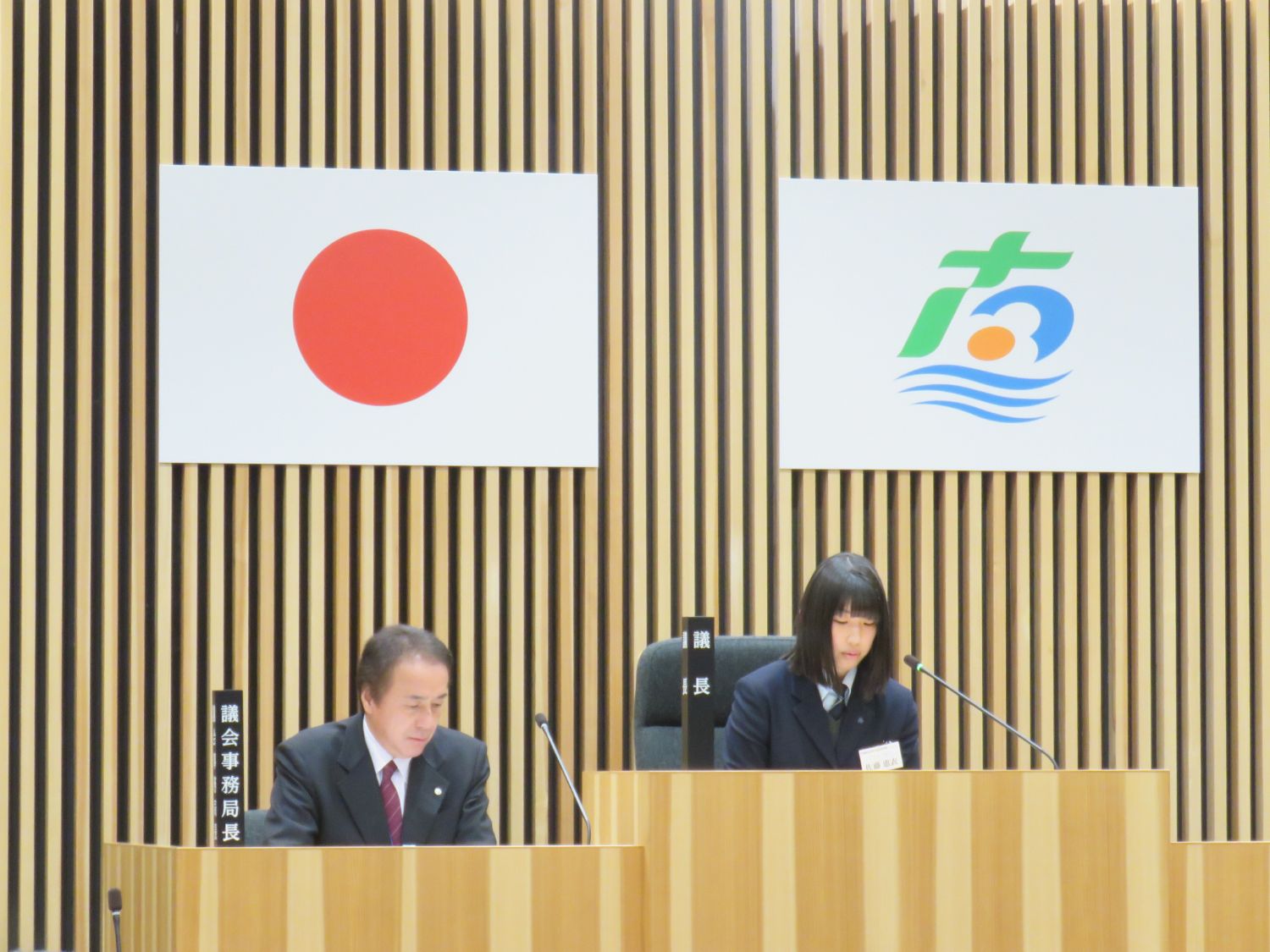 国旗と町旗が掲げられている壁の前に設置された議長席に佐藤恵衣さんが座り、話をしている様子の写真