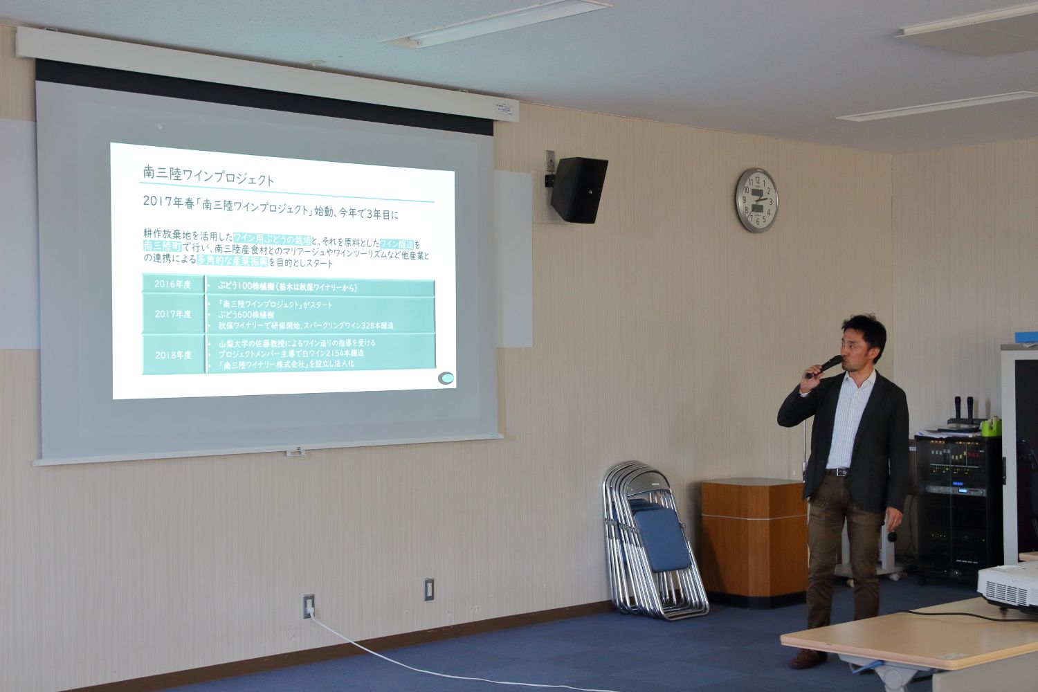 「南三陸ワインプロジェクト」についての資料が映し出されたスクリーンが設置された会議室で、右側に立っている男性がマイクを使って話をしている様子の写真