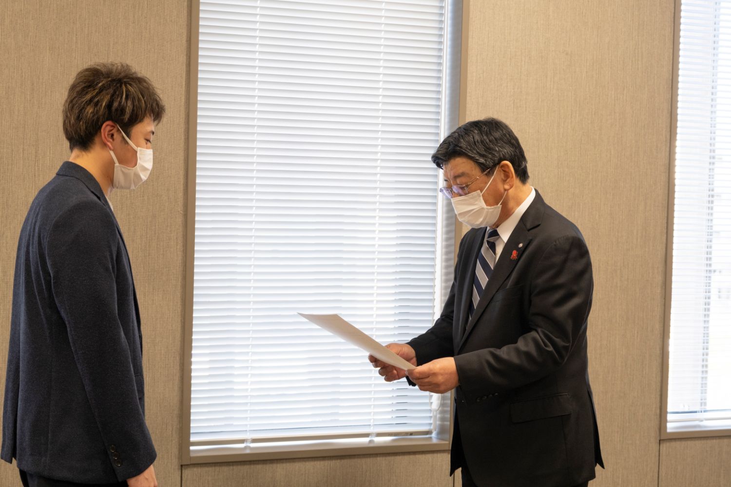 紺色のジャケットと白いマスクをつけた佐藤さんが、黒いスーツにストライプのネクタイを締めた南三陸町長と向かい合い、南三陸町長が委嘱状を読み上げているのを聞いている写真