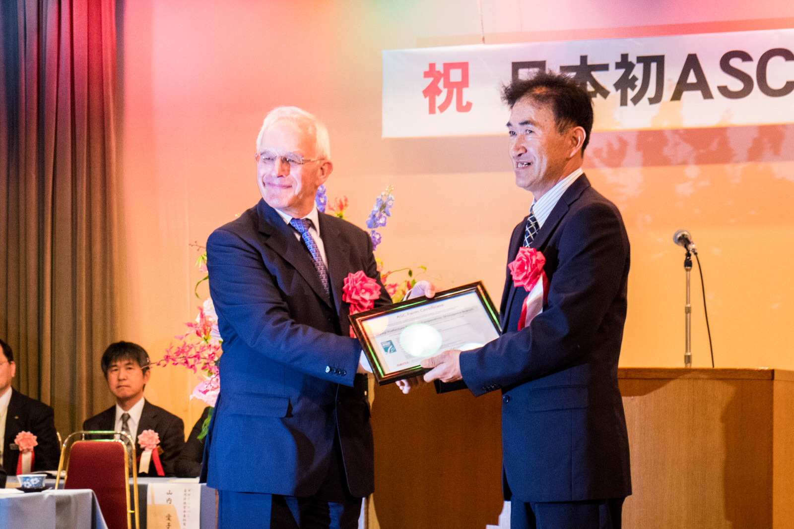 南三陸町が日本で初めてASC認証を取得したことを記念する伝達式表彰式で、スーツを着た二人の男性が壇上で賞状を笑顔で受け渡している様子を写した写真