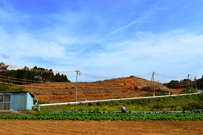 畑の奥にショベルカーが作業する造成中の土の斜面があり、手前に農作業中の人とビニールハウスが見える高台移転中の写真