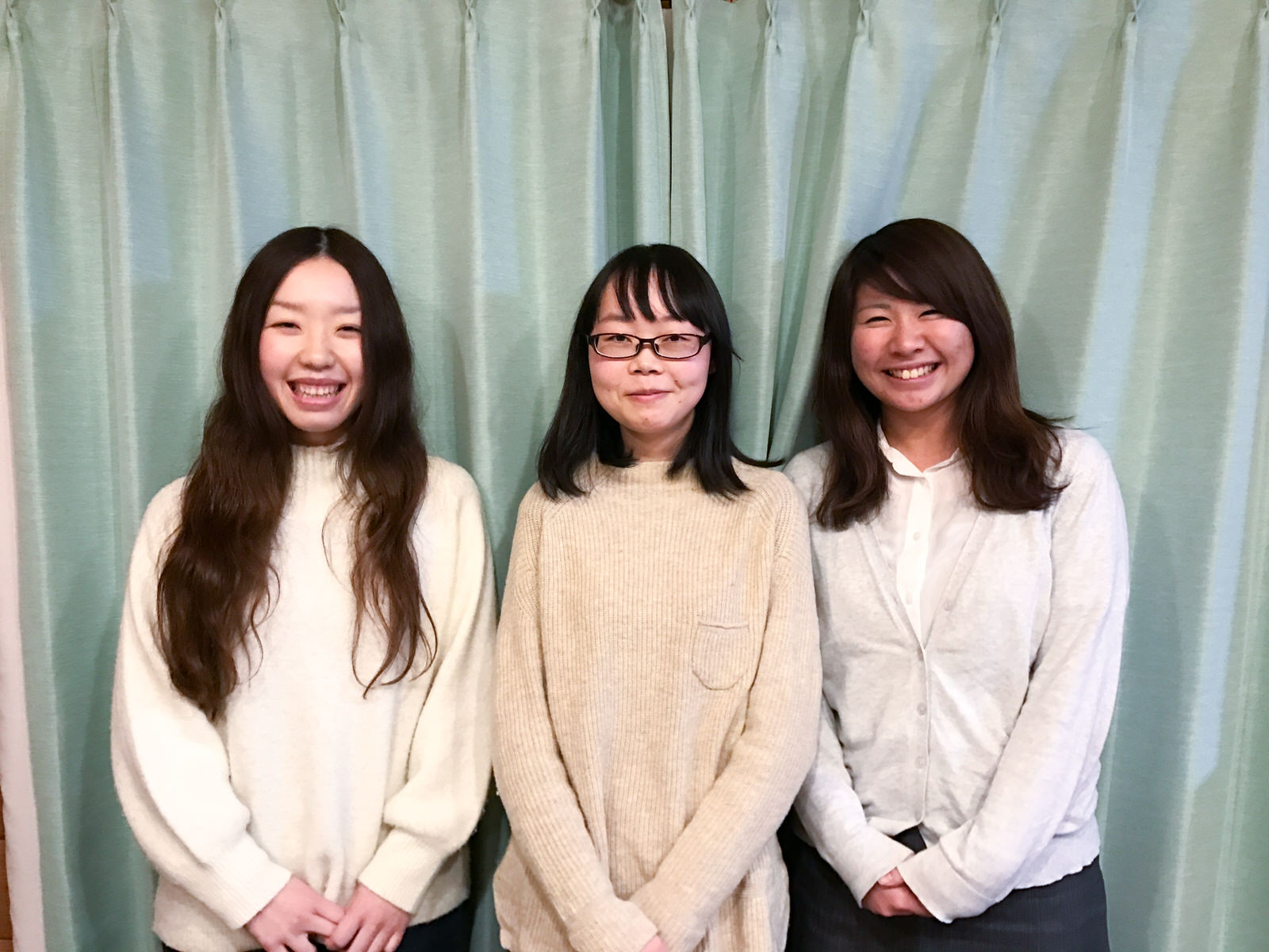水色のカーテンの前で3人の女性が笑顔で並んで立っている写真