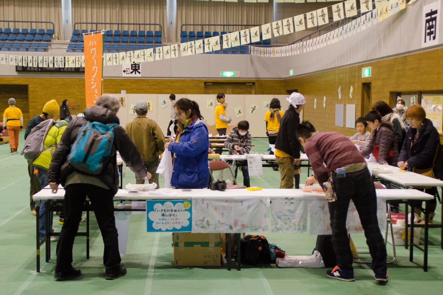 壁や天井に鳥の絵が飾られた会場内に、いくつものテーブルが並び、子どもから大人までの参加者がスタッフと一緒に工作などのワークショップを楽しんでいるイベント会場の様子の写真