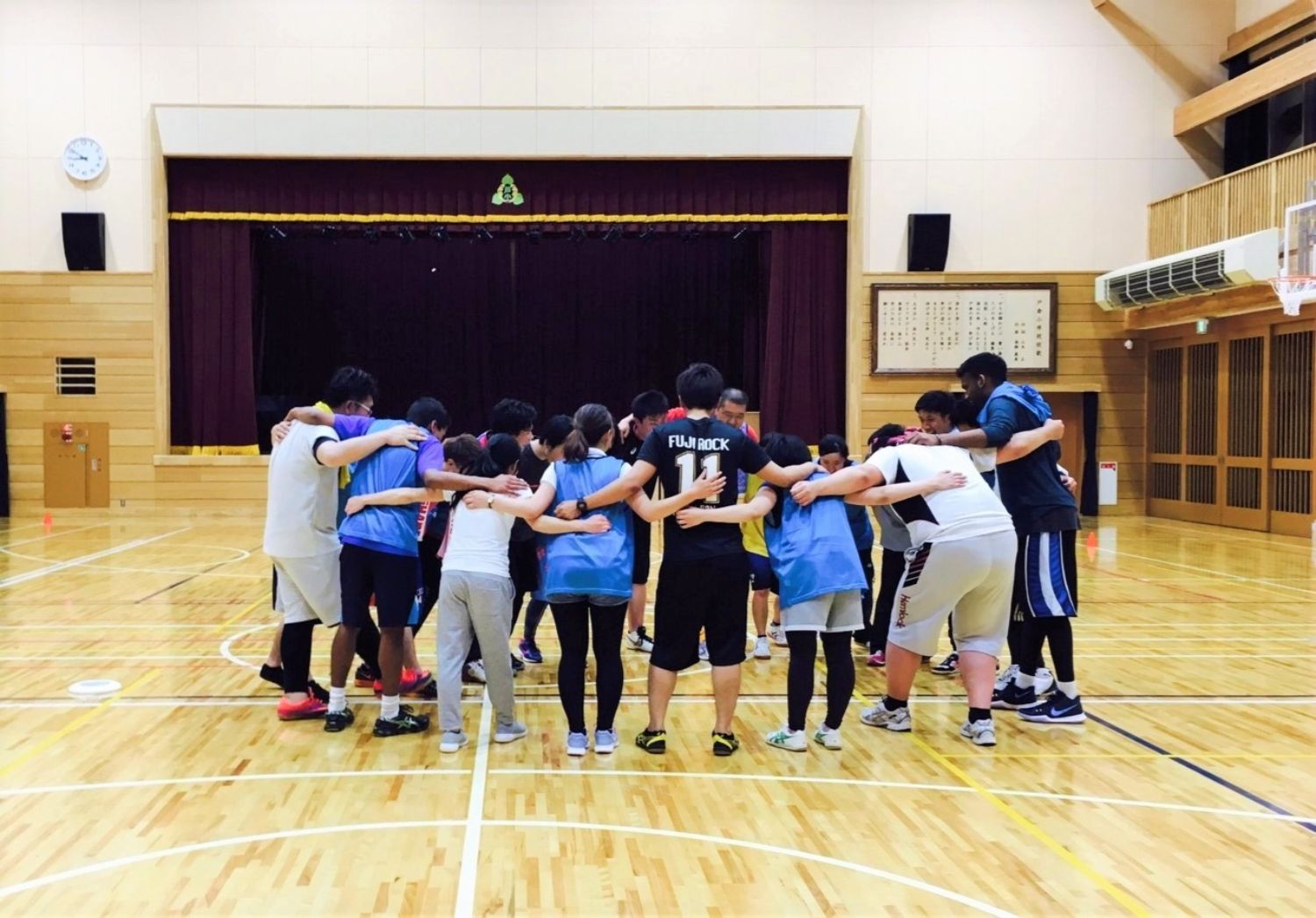 床は磨かれた木製で、背景にはステージが見える体育館の中で、複数の人物が円になって肩を組み合って立っている様子を写している写真