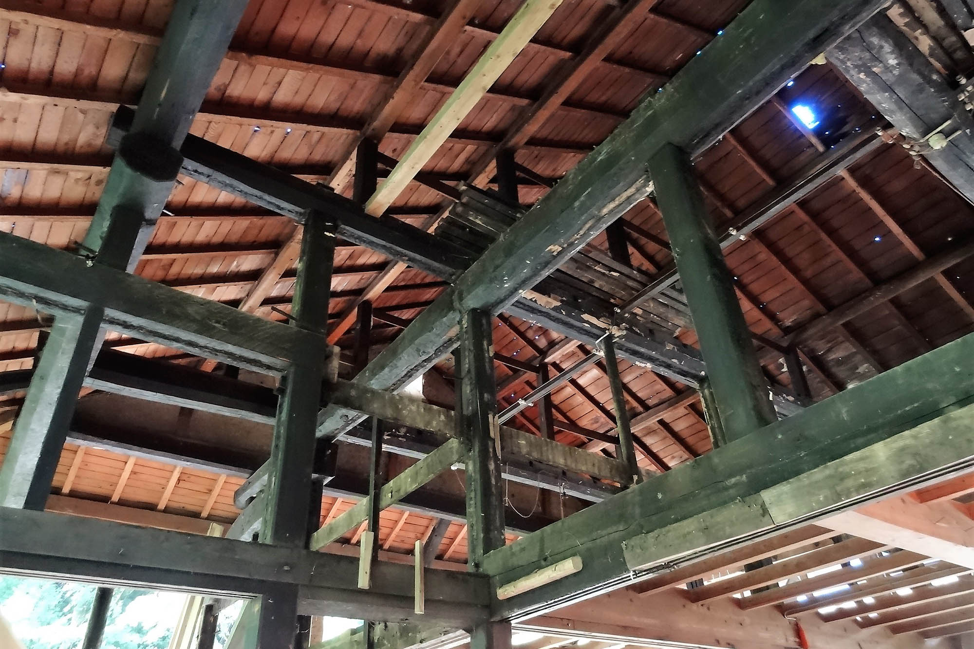 古民家の梁や柱がむき出しになった状態で、赤茶色の屋根裏が見える開放的な空間に、太い黒い木材が交差し、修繕や補強の跡が随所に見られるリノベーション中の古民家内部の写真