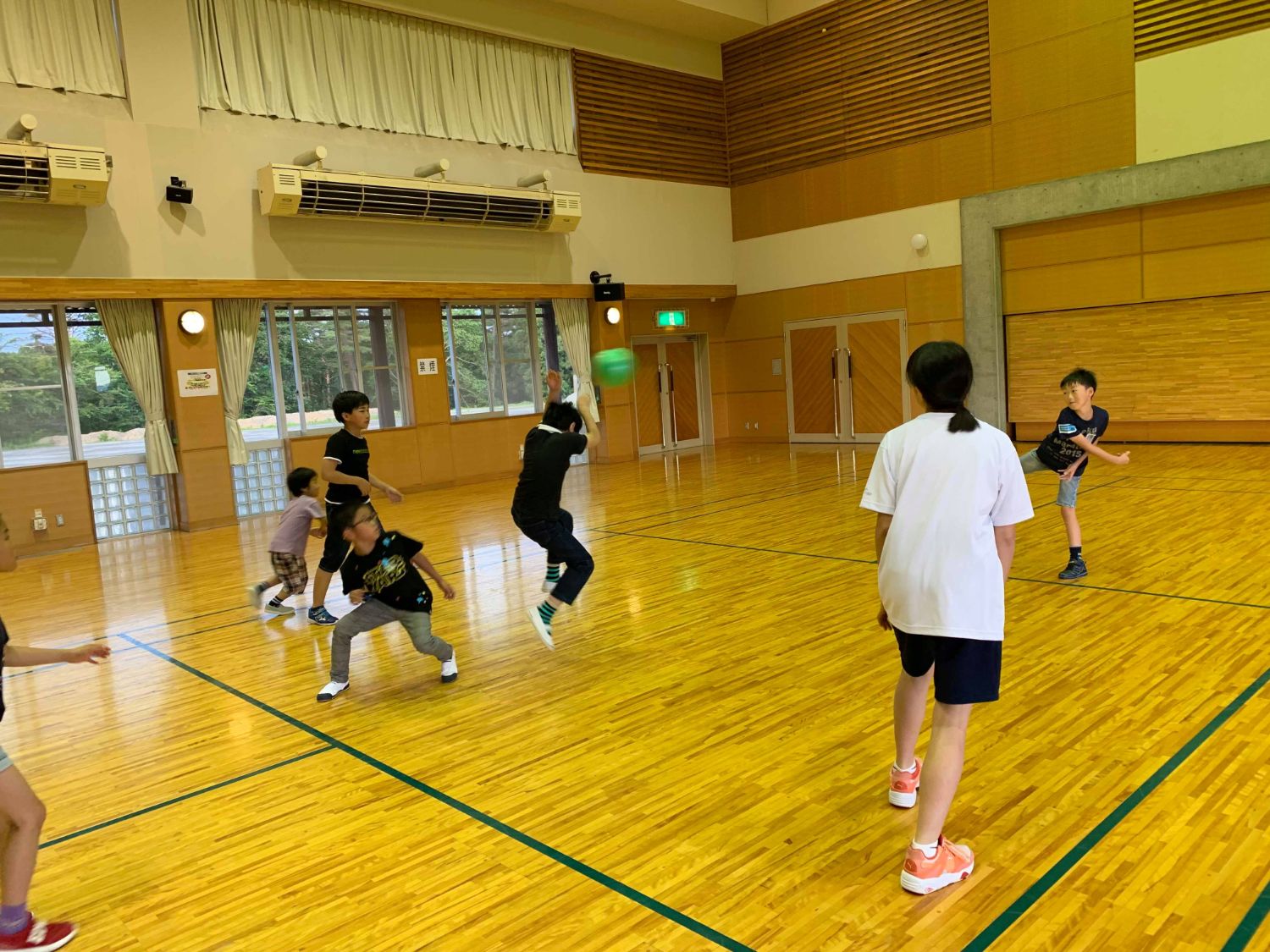 体育館内で子供たちがドッジボールを行い、右側の男の子が黒いシャツを着た子どもに投げた緑色のボールが当たっている写真