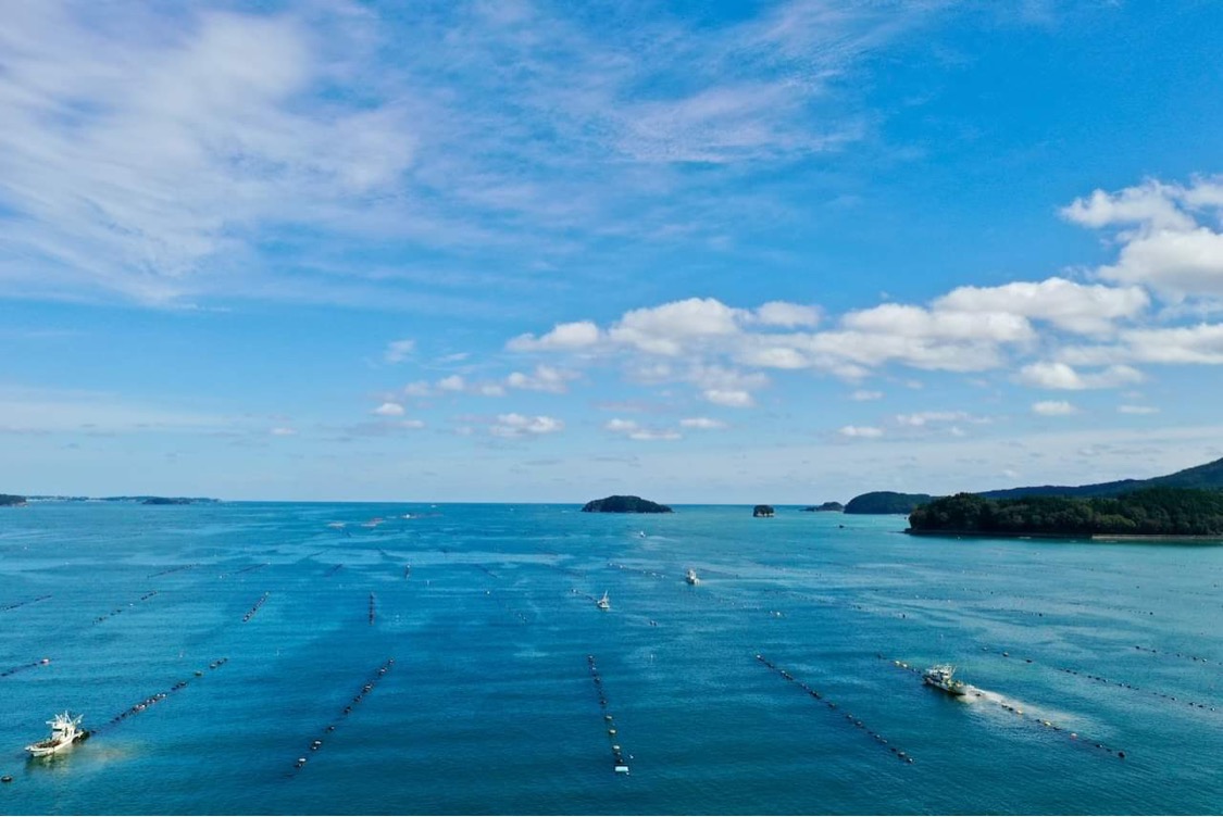 雲が浮かぶ青空のもと、海には多数の養殖いかだが浮かび、数隻の漁船が航行している様子を捉えた写真