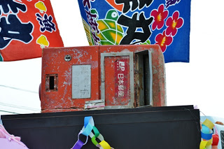 カラフルなのぼり旗の前に、津波被害を受けて錆びて傷だらけの赤い郵便ポストが台の上に飾られている写真