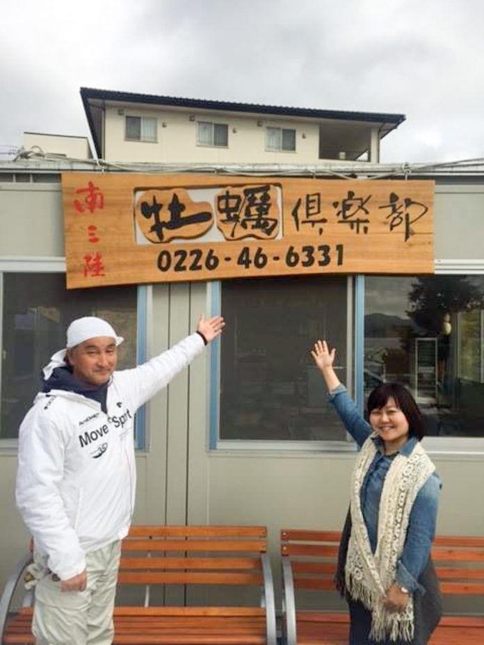 コンテナハウスに設置された「南三陸 牡蠣倶楽部」と書かれた木製の看板を手で示している頭にタオルを巻いた男性と女性リポーターの写真