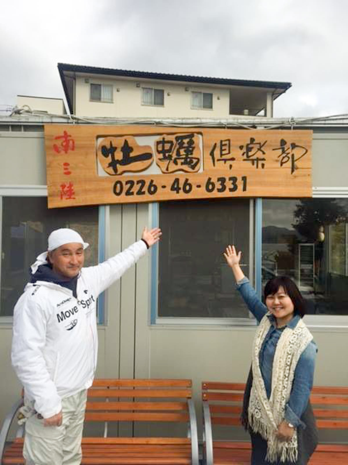 コンテナハウスに設置された「南三陸 牡蠣倶楽部」と書かれた木製の看板を手で示している頭にタオルを巻いた男性と女性リポーターの写真